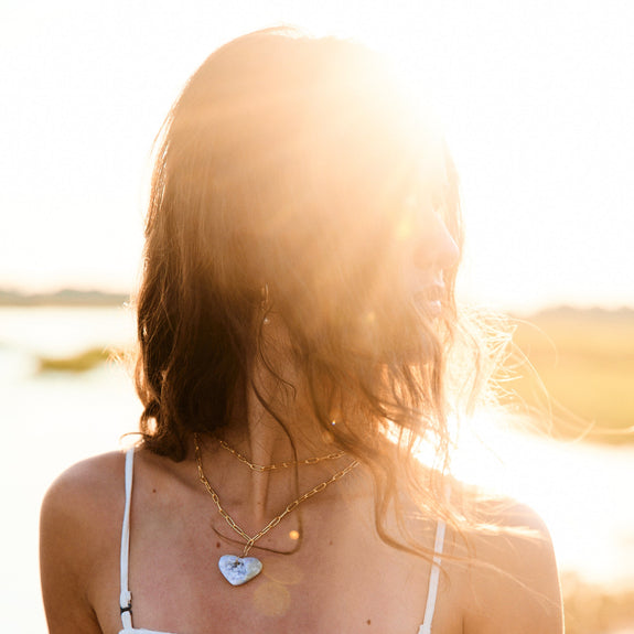 Woman in a white dress with layered necklaces standing in front of a sunset.
